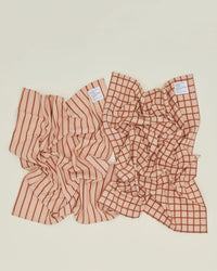 Two Essential Dish Towels in Blush/Terracotta by Hawkins New York rest on a light surface; one features vertical stripes, the other a grid pattern, both in beige and brown tones.