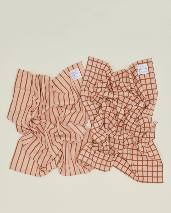 Two Essential Dish Towels in Blush/Terracotta by Hawkins New York rest on a light surface; one features vertical stripes, the other a grid pattern, both in beige and brown tones.