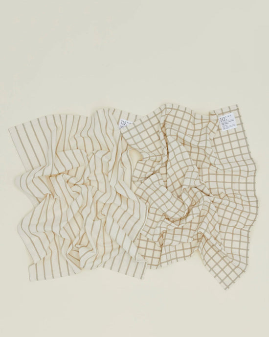 The Hawkins New York Essential Dish Towels in Flax/Ivory (Set of 2) are shown on a light surface; crafted from Oeko-Tex cotton, one towel features vertical stripes and the other a grid pattern.