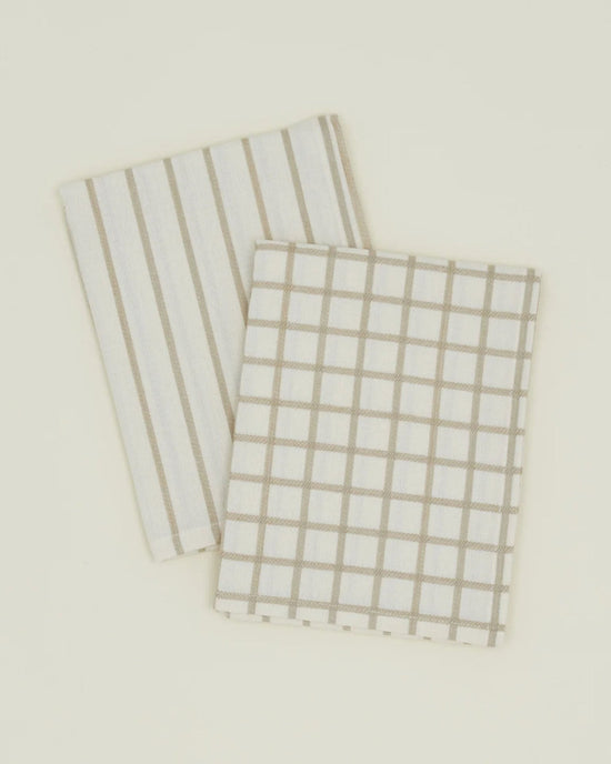 A pair of Hawkins New York Essential Dish Towels in Flax/Ivory—one striped, one grid patterned—crafted from Oeko-Tex cotton, displayed folded on a light surface.