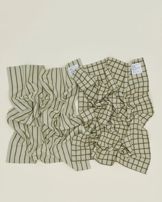 Set of two olive and sage Essential Dish Towels by Hawkins New York, each with a unique pattern—one striped, one grid—and white labels in the top right corner. Perfect absorbent cotton towels for any kitchen.