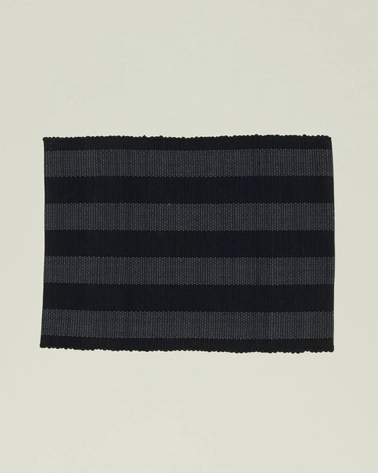 The Essential Floor Mat in Black by Hawkins New York features alternating black and dark gray stripes, is rectangular, crafted from recycled fiber, fully machine washable, and displayed on a plain light background.
