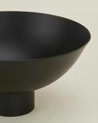 The Essential Footed Bowl in Black by Hawkins New York features a matte black, minimalist design with a wide, rounded shape on a short cylindrical base, displayed against a plain light background.