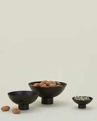 Three black, modern pedestal bowls are arranged on a light surface. The largest and smallest serve nuts, while the medium bowl is empty with walnuts beside it. One is the Essential Footed Bowl in Black by Hawkins New York; a beige background adds warmth.