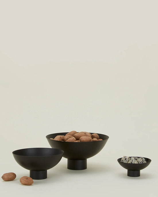 Three black, modern pedestal bowls are arranged on a light surface. The largest and smallest serve nuts, while the medium bowl is empty with walnuts beside it. One is the Essential Footed Bowl in Black by Hawkins New York; a beige background adds warmth.