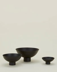 Three Hawkins New York Essential Footed Bowls in matte black, each with a wide, shallow shape and short cylindrical base, are arranged in a row against a plain light beige background.