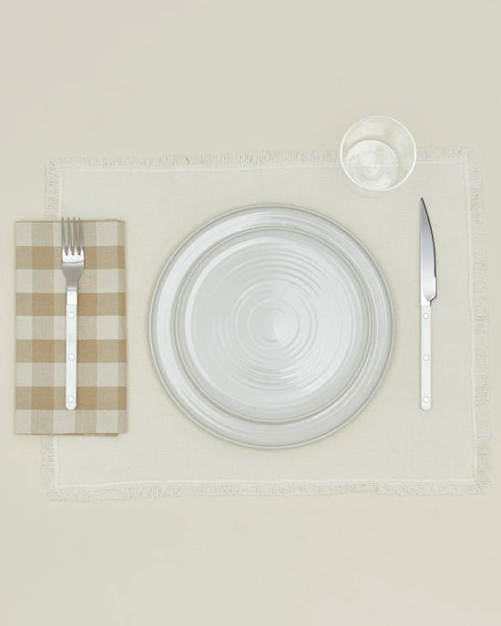 A table setting with a white plate, glass, and fork on Hawkins New York Essential Gingham Dinner Napkins in Ivory/Flax—set of 4—to the left, knife to the right, all neatly arranged on a beige placemat.