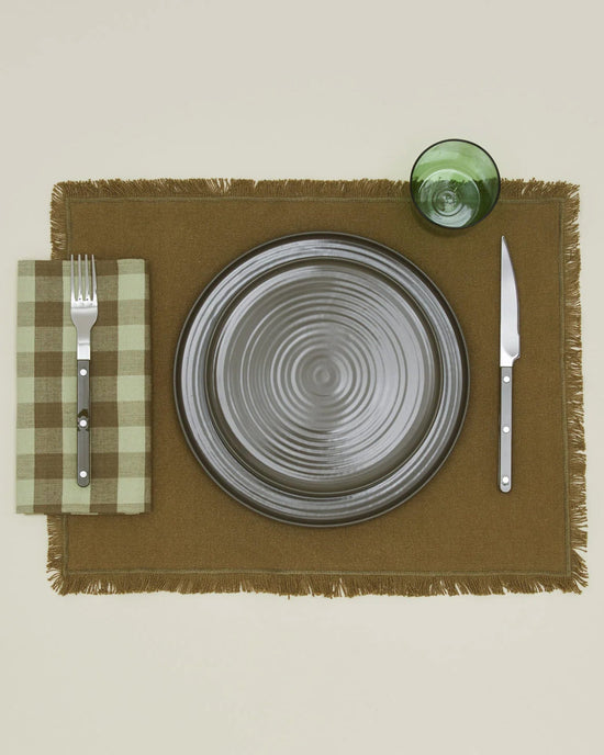 A gray ceramic plate sits on a brown placemat, flanked by a fork on Hawkins New Yorks Essential Gingham Dinner Napkins in Sage/Olive to the left, a knife to the right, and a green glass above.