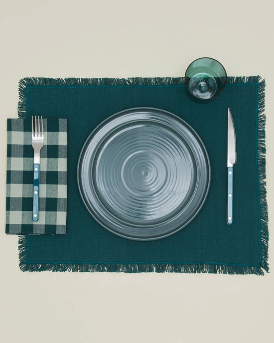 A table setting with a gray plate, fork on a Hawkins New York Essential Gingham Dinner Napkin in Sky/Peacock, knife, green glass, all arranged on a dark green fringed placemat.