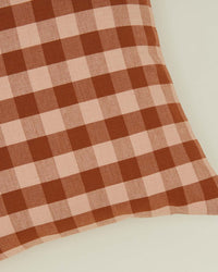 A close-up of the Essential Gingham Pillow (18x18 inches, Blush/Terracotta) by Hawkins New York, featuring a brown and pink checkered pattern on a light surface.