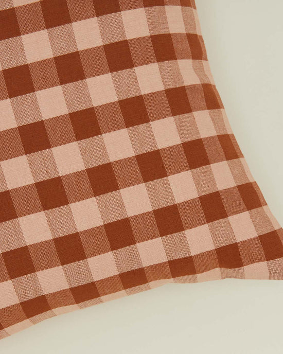 A close-up of the Essential Gingham Pillow (18x18 inches, Blush/Terracotta) by Hawkins New York, featuring a brown and pink checkered pattern on a light surface.