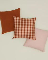 Three square throw pillows are arranged on a light surface: a solid rust Hawkins New York European linen pillow with down insert, the 18x18-inch Essential Gingham Pillow in Blush/Terracotta by Hawkins New York, and a solid light pink pillow.