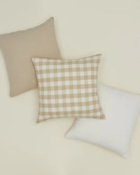 Three square pillows sit on a light surface: one beige, one white, and the Hawkins New York Essential Gingham Pillow (18x18, Ivory/Flax) featuring a beige and white checkered pattern.