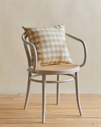 A light wooden chair with curved arms and a woven seat displays the Hawkins New York Essential Gingham Pillow (18x18, Ivory/Flax) on a wooden floor, set against a plain background.