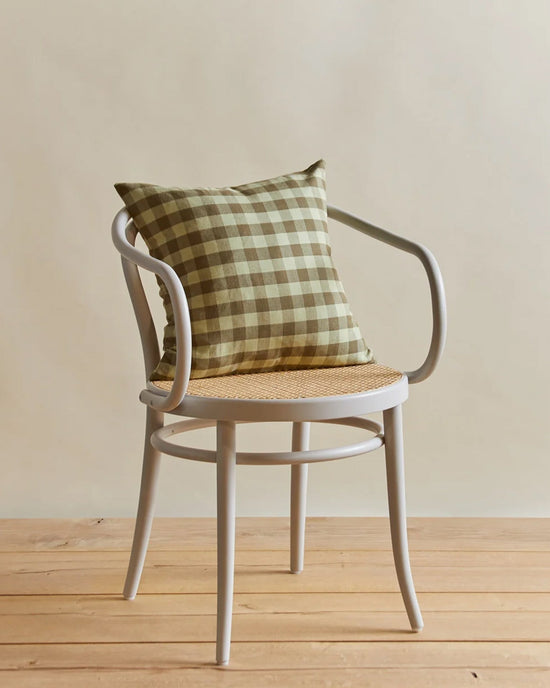 A white wooden chair sits on a wooden floor with the Hawkins New York Essential Gingham Pillow in Sage/Olive (18x18 inches), featuring a plush down insert, set against a neutral background.