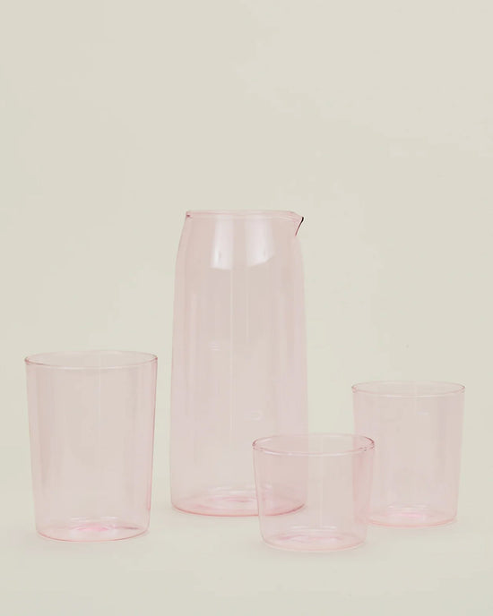 A Hawkins New York Essential Glassware - Pitcher in Blush and three matching translucent pink cups are displayed against a plain, light background.