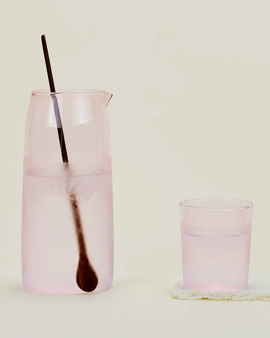 A Hawkins New York Essential Glassware - Pitcher in Blush stands beside a matching pink glass, both filled with water and accompanied by a long metal spoon, all set against a plain background.