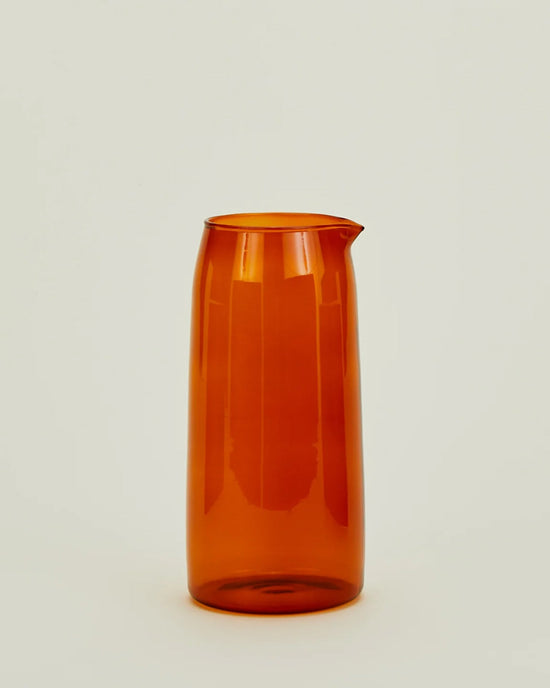 The Hawkins New York Essential Glassware Pitcher in Terracotta, crafted from translucent orange borosilicate glass and featuring a spout, is centered against a plain off-white background.
