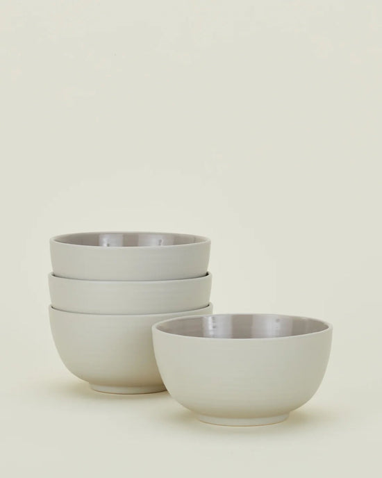 Four Essential Large Bowls in Light Grey by Hawkins New York, three stacked on the left and one apart on the right, are displayed against a light background. This set of dishwasher-safe stoneware combines elegance with everyday ease.