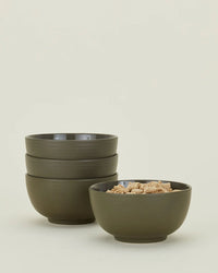 Four Essential Large Bowls in Olive by Hawkins New York—three stacked, one filled with square cereal—are displayed on a plain, light backdrop. This stoneware dinnerware set of 4 is dishwasher safe.