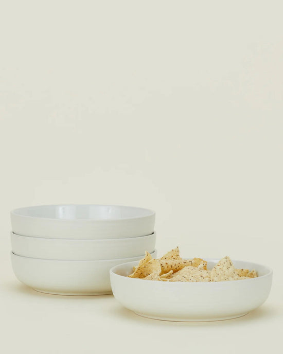 Four Hawkins New York Essential Low Bowls in Bone are stacked on the left, with one matching bowl filled with tortilla chips on the right—versatile stoneware dinnerware ideal for any occasion, set against a simple light background.