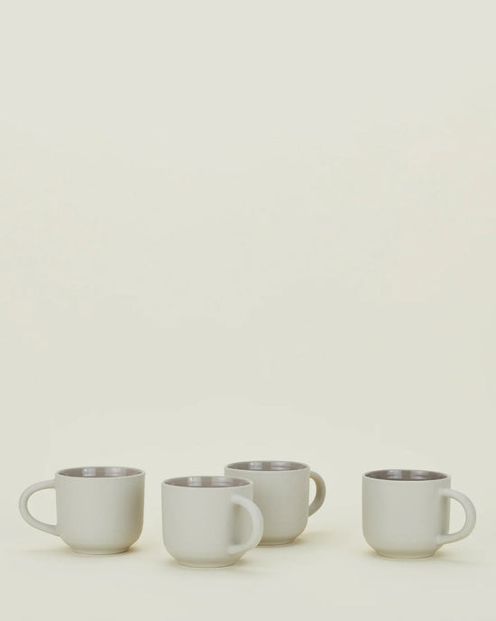 Four light grey stoneware mugs with handles from Hawkins New Yorks Essential Mugs set are arranged in a row on a light surface against an off-white background—timeless, elegant, and dishwasher safe.
