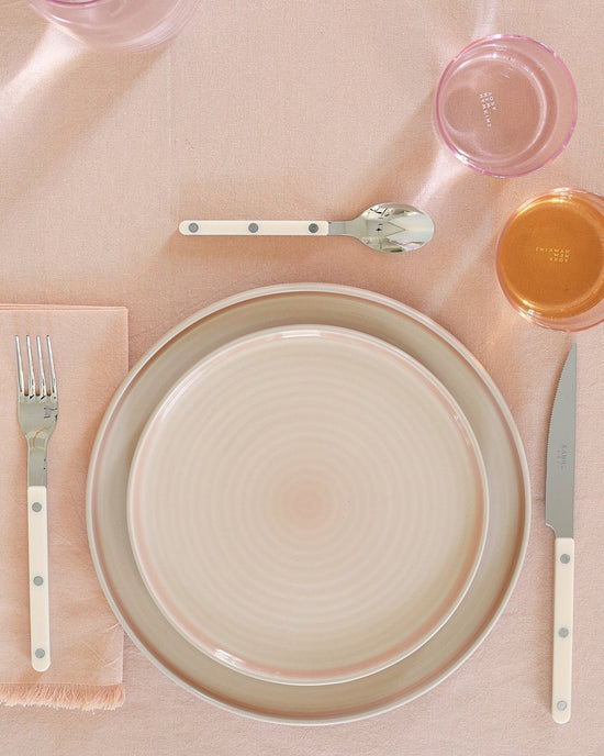 A neatly arranged table features a fork, knife, spoon, Hawkins New York Essential Salad Plates in Blush (set of 4), and two pink-tinted glasses placed on a light pink tablecloth.