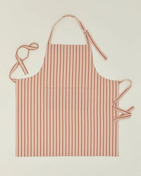 The Hawkins New York Essential Striped Apron in Blush/Terracotta is a cotton apron featuring vertical stripes, an adjustable halter neck loop, two waist ties, and a large central pocket, shown laid flat on a plain background.