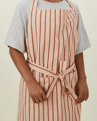 A person wears a light grey T-shirt and the Hawkins New York Essential Striped Apron in Blush/Terracotta, featuring vertical stripes and an adjustable halter, tied at the waist, against a plain background.