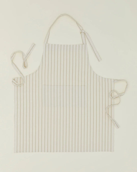 The Essential Striped Apron in Ivory/Flax by Hawkins New York is a cotton apron with vertical beige stripes and two front pockets, shown flat with ties extended—a stylish kitchen gift for any home chef.
