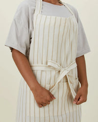Someone wears the Hawkins New York Essential Striped Apron in Ivory/Flax—crafted in cotton with vertical stripes, front pockets, and a tied waist—over a short-sleeved shirt, making it an ideal kitchen gift.