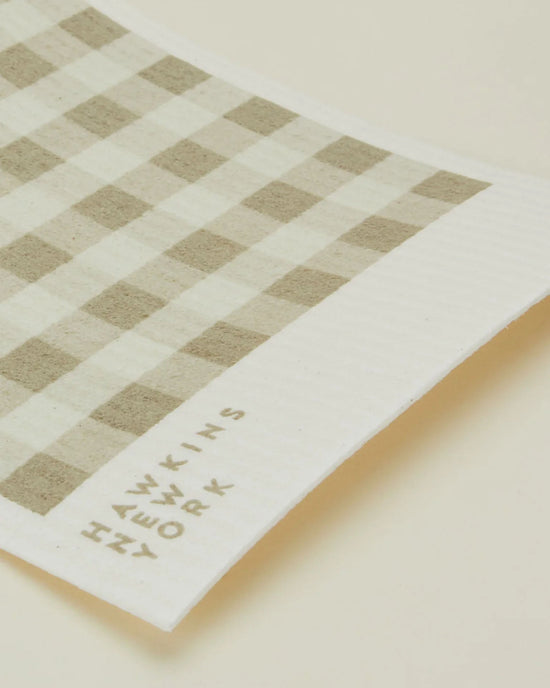 A close-up of Hawkins New York’s Essential Swedish Dishcloths in Ivory/Flax (set of 6), featuring a beige and white check pattern with the brands name printed in the corner.