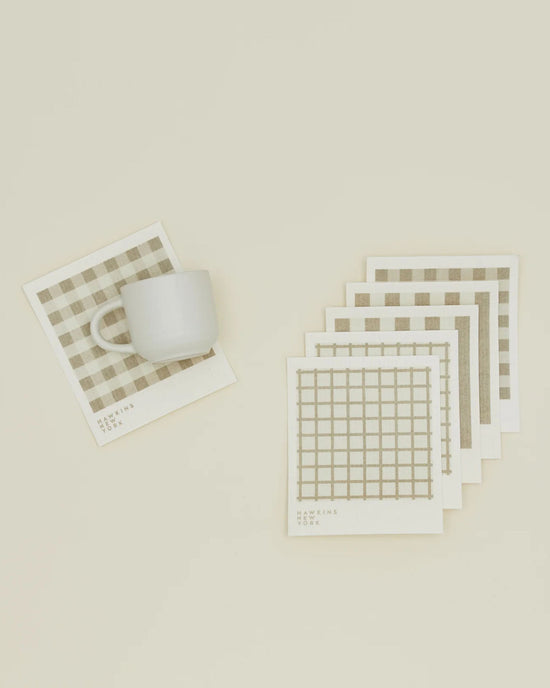 A white ceramic mug sits on an Essential Swedish Dishcloth by Hawkins New York in Ivory/Flax, with five more grid-patterned dishcloths from the set of 6 arranged nearby on a cream surface.