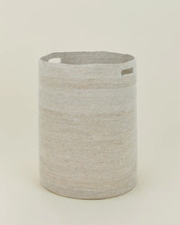 The Essential Woven Hamper by Hawkins New York is a round, light beige handwoven seagrass hamper with two rectangular cutout handles, shown upright against a plain off-white background.