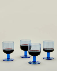 Four blue-stemmed wine glasses from the Hawkins New York Set of 4 Stemmed Simple Seeded Glassware, each half-filled with red wine, are arranged on a light surface against a plain beige background.