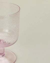 A close-up of a translucent blush Hawkins New York Set of 4 Stemmed Simple Seeded Glassware, featuring small bubbles in the glass, set against a plain light background.