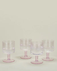 A set of 4 Stemmed Simple Seeded Glassware in Blush by Hawkins New York, partially filled with water and ice, is arranged on a light surface against a plain background.