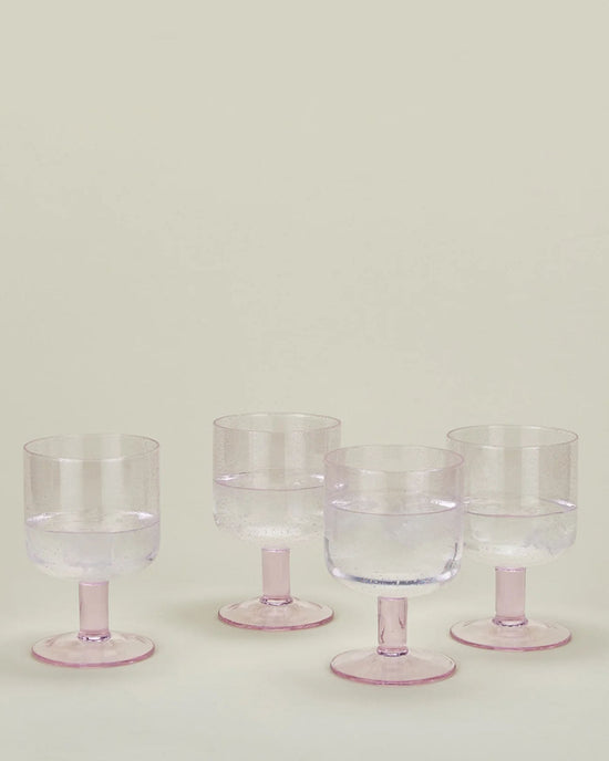 A set of 4 Stemmed Simple Seeded Glassware in Blush by Hawkins New York, partially filled with water and ice, is arranged on a light surface against a plain background.
