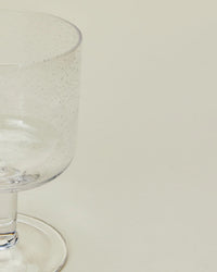A partially shown Hawkins New York Set of 4 Stemmed Simple Seeded Glassware in Clear, featuring small bubbles, displayed against a plain light background.