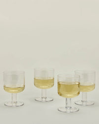 A Set of 4 Stemmed Simple Seeded Glassware in Clear by Hawkins New York, each partially filled with pale yellow liquid, is arranged on a light surface against a neutral background.