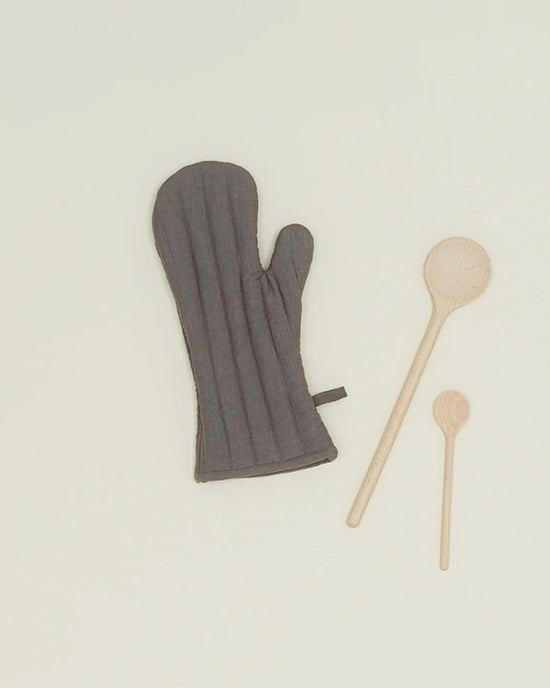 A Hawkins New York Simple Linen Oven Mitt in Dark Grey with heat-resistant padding is placed beside two wooden spoons, one large and one small, on a plain light background.