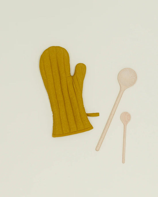 The Hawkins New York Simple Linen Oven Mitt in Mustard sits beside two differently sized wooden spoons on a light cream background.