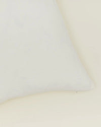 A close-up of the bottom right corner of the Hawkins New York Simple Linen Pillow—18 x 18 inches in ivory with a down insert—set against a light beige background.
