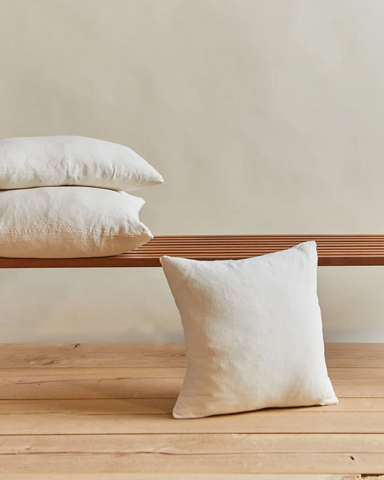 Two Hawkins New York Simple Linen Pillows in ivory rest on a slatted wooden bench, while a third 18 x 18-inch pillow sits on the light wood floor against a beige wall in this serene, minimalist space.