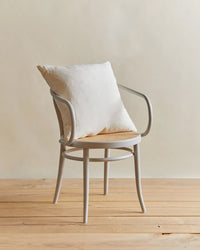 A light gray wooden chair with curved armrests and a round woven seat holds the Hawkins New York Simple Linen Pillow (22 x 22 inches, Ivory), featuring a plush down insert, on a wooden floor against a neutral beige background.