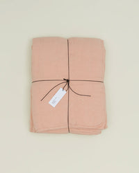 A folded Hawkins New York Simple Linen Quilt in Blush, tied with dark string and a white tag, rests on a plain light background.