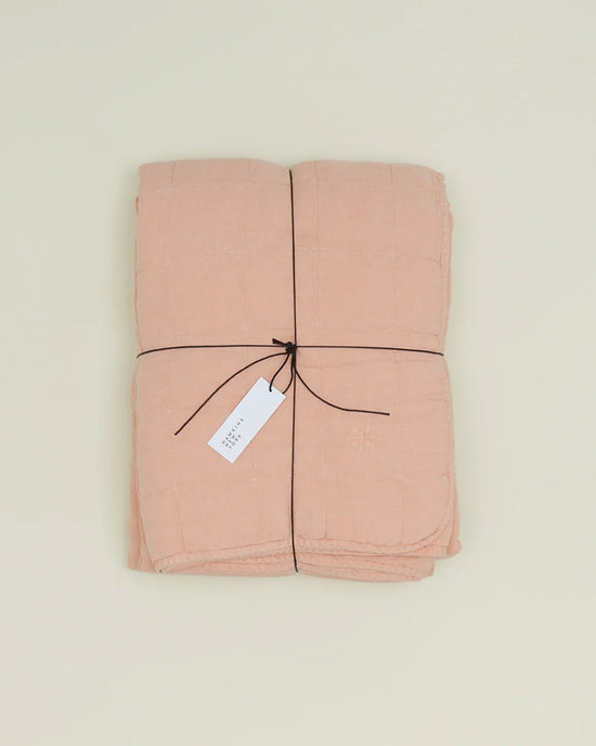 A folded Hawkins New York Simple Linen Quilt in Blush, tied with dark string and a white tag, rests on a plain light background.