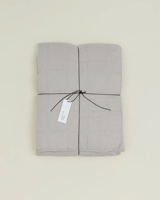 The Simple Linen Quilt in Light Grey by Hawkins New York, stonewashed and folded, is tied with a black string and a white tag, set on a plain light background.