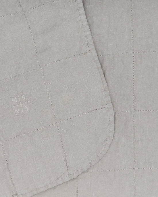Close-up of the Simple Linen Quilt in Light Grey by Hawkins New York, featuring a stitched grid pattern and an embroidered HN NY logo on one corner.