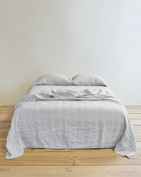 A neatly made bed features the Hawkins New York Simple Linen Quilt in Light Grey and two pillows, resting on a wooden floor against a plain white wall.